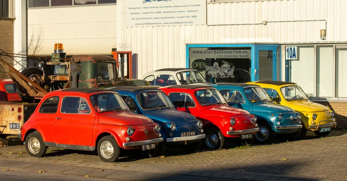 Rangée de voitures Fiat 500 vintage colorées garées au soleil devant un garage.