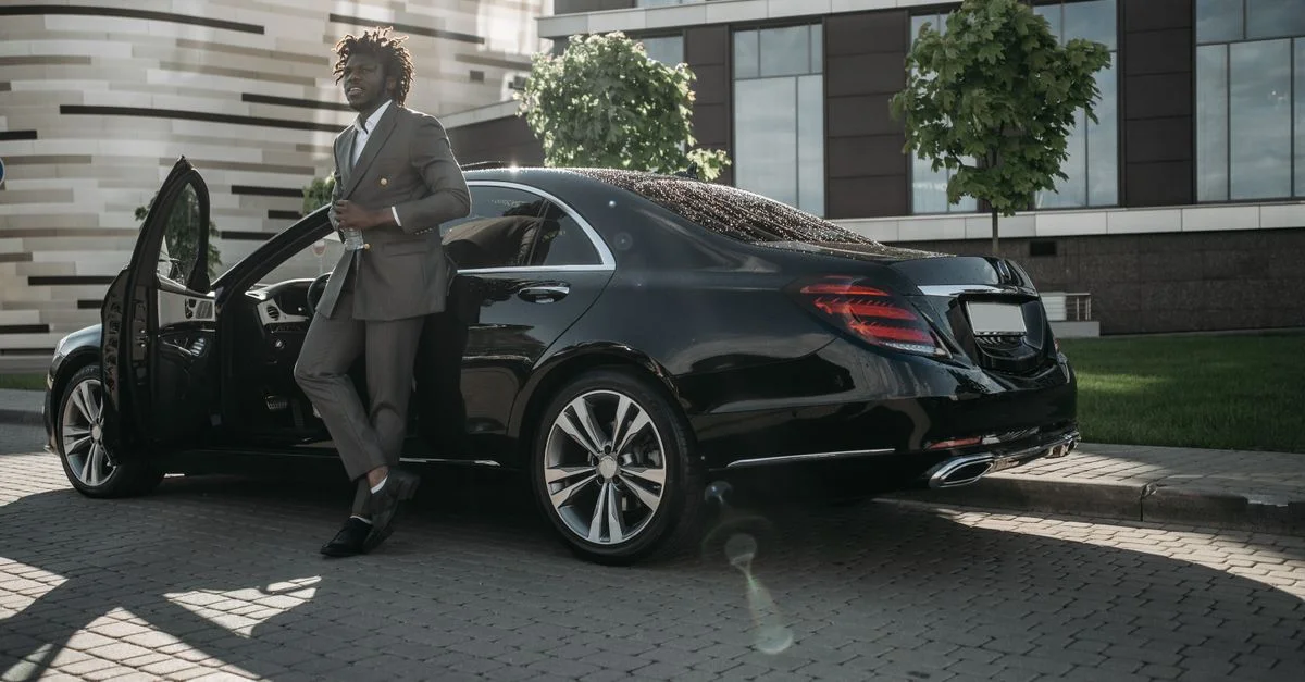 Un homme d'affaires en costume gris avec des dreadlocks sortant d'une voiture de luxe noire dans un cadre urbain.