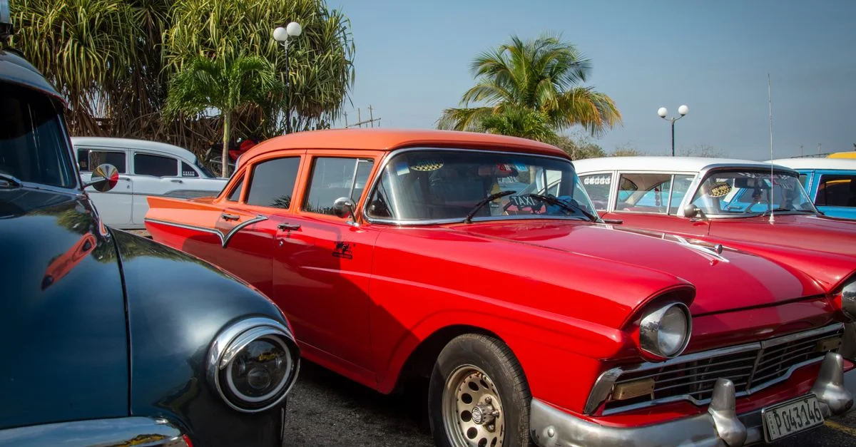 Une collection de voitures classiques vintage, dont une Ford rouge, dans un parking extérieur sous un ciel bleu clair.