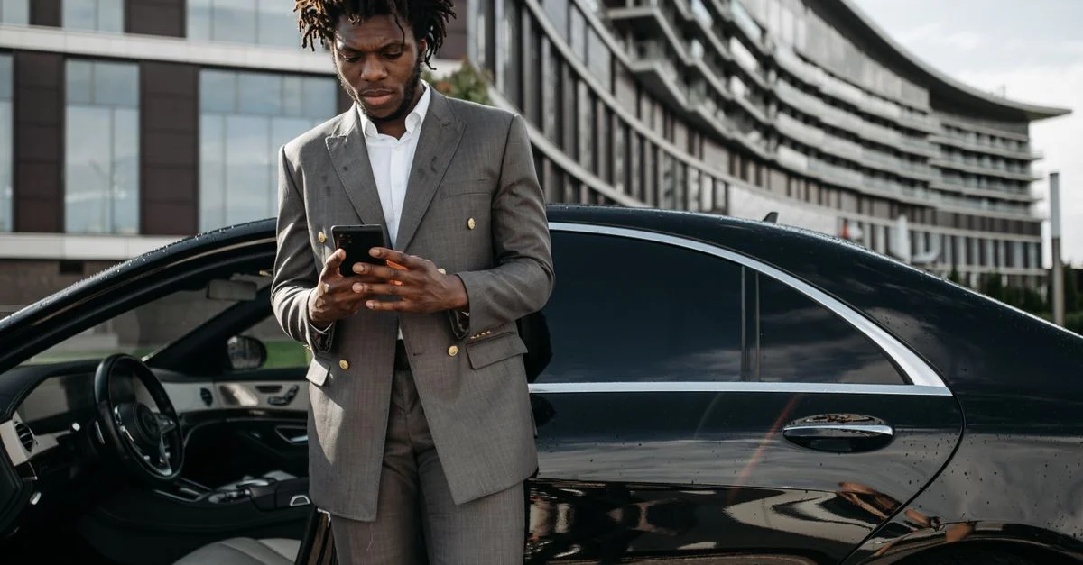 Homme professionnel en costume gris utilisant son téléphone à côté d'une voiture de luxe.